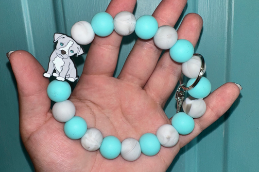 Hand holding a bracelet with turquoise and white beads and a dog charm against a teal background