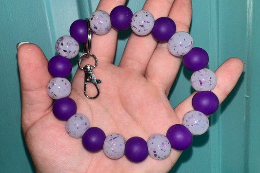 Hand holding a purple and gray beaded keychain against a teal background